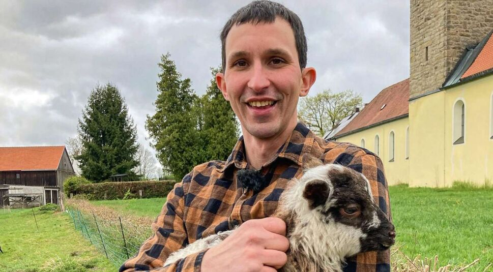 Bei Bauer sucht Frau 2025 hält Kandidat Michael ein schwarz-weißes Lämmchen im Arm. Mit kariertem Hemd steht er lächelnd auf einer grünen Wiese vor ländlicher Kulisse mit Kirche und Hof.