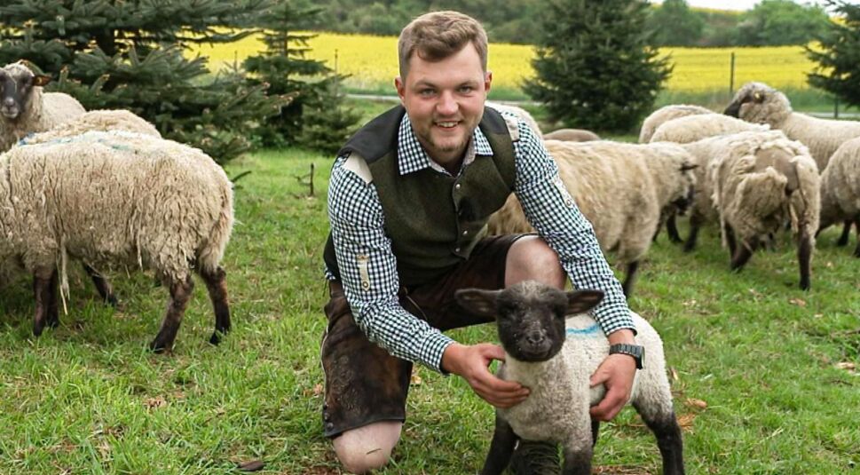 Bei Bauer sucht Frau 2025 kniet Kandidat Michi im traditionellen Outfit auf einer Schafweide und hält ein junges Lämmchen. Er lächelt herzlich und wirkt naturverbunden und bodenständig.