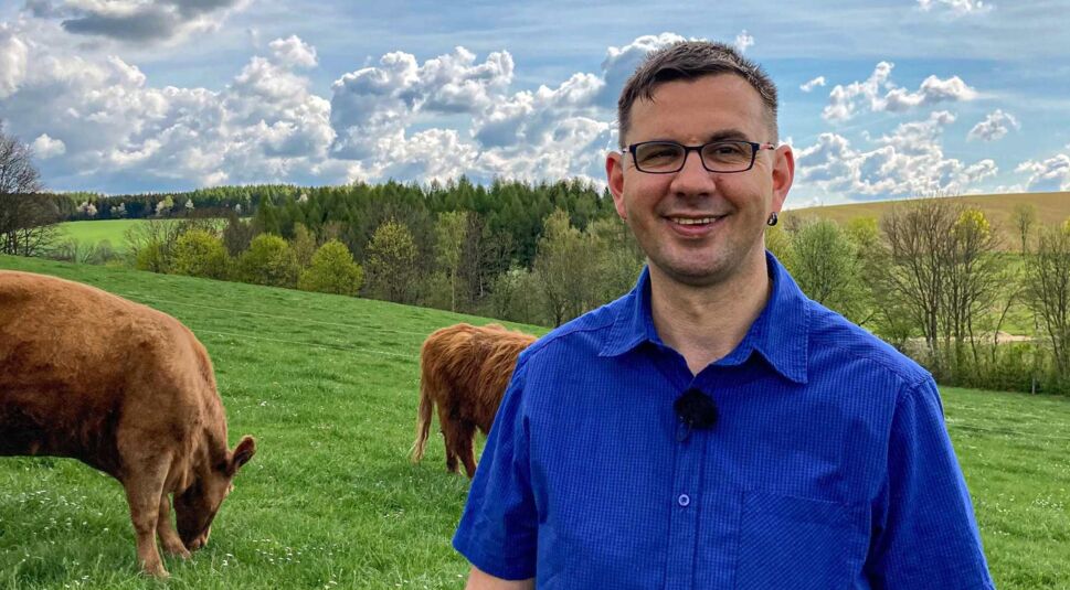 Bei Bauer sucht Frau 2025 steht Kandidat Thomas in blauem Hemd auf einer grünen Weide mit grasenden Rindern. Im Hintergrund erstreckt sich eine hügelige Landschaft unter einem dramatischen Himmel.