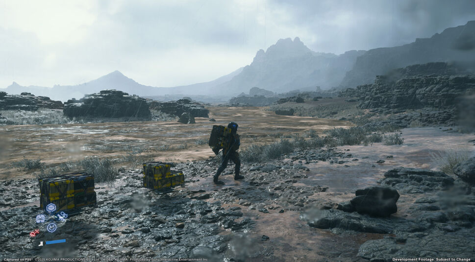 In diesem Screenshot aus Death Stranding 2: On the Beach durchquert ein einsamer Bote eine karge, regennasse Felslandschaft. Auf seinem Rücken trägt er schwere Ausrüstung, während weitere Fracht an einem Seil hinter ihm hergezogen wird. Die düstere Umgebung mit Bergen und Sturm vermittelt eine bedrückende Atmosphäre.