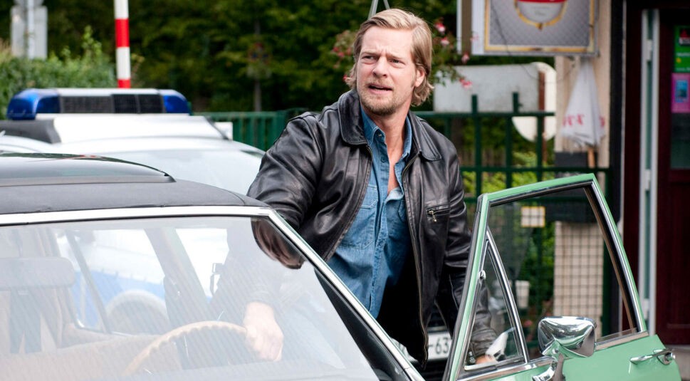 Auf dem Bild zur Serie Der letzte Bulle ist ein blonder Mann mit Bart zu sehen, der eine schwarze Lederjacke und ein offenes Jeanshemd trägt. Er steigt gerade aus einem mintgrünen Oldtimer mit geöffneter Fahrertür. Im Hintergrund steht ein Polizeiwagen.