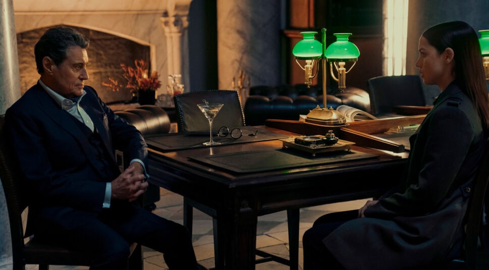 Im Bild zur John Wick Ballerina Kritik sitzen Ian McShane als Winston Scott und Ana de Armas als Eve sich in einem eleganten, dunklen Raum gegenüber. Winston trägt einen Anzug und hat die Hände gefaltet, während Eve ernst zuhört. Auf dem Tisch stehen eine grüne Bankerlampe, ein Martini-Glas und eine Brille, was die gehobene Atmosphäre unterstreicht.