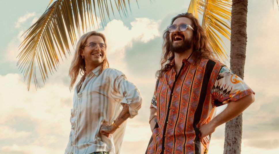 Auf dem symbolischen Bild zur Kaulitz & Kaulitz Staffel 3 stehen Bill und Tom Kaulitz entspannt unter Palmen im Sonnenuntergang. Bill trägt ein hell gestreiftes Hemd und blickt lächelnd zu seinem Bruder, der mit farbenfrohem Musterhemd herzhaft lacht. Die Stimmung ist unbeschwert und sommerlich – ein Hinweis auf mögliche neue Folgen.