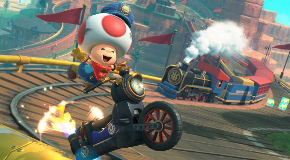 Auf dem Bild zu Mario Kart World Tipps springt Toad in einer Lokführer-Uniform fröhlich von seinem Motorrad ab, während im Hintergrund eine bunte Dampflok durch eine kurvige Bergstrecke fährt. Die Szene ist voller Bewegung, mit glühenden Reifen, Schienen und Items – sie vermittelt Abenteuerlust und den spaßigen Chaos-Faktor von Mario Kart.