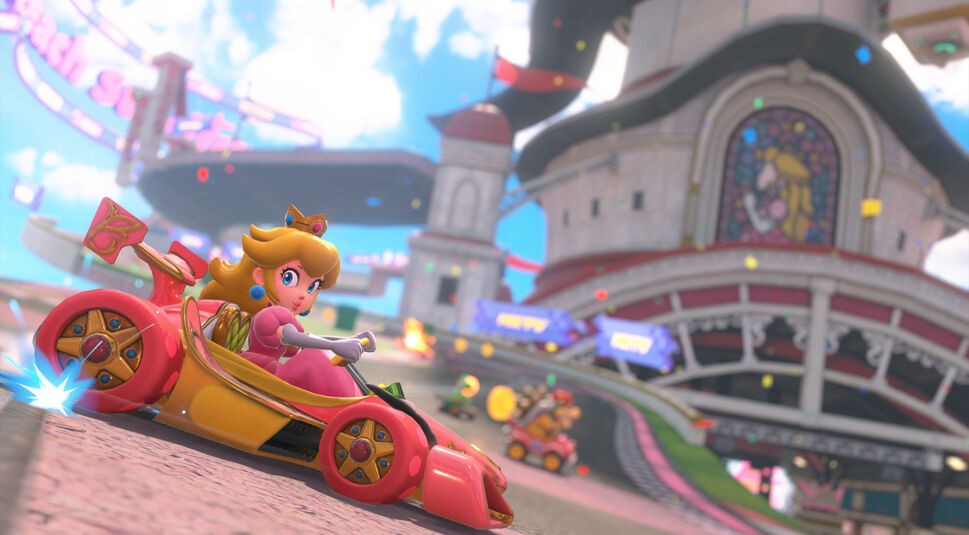 Das Bild in dem Artikel zum Mario Kart World-Test zeigt Prinzessin Peach in einem rosafarbenen Kart auf einer königlichen Rennstrecke. Im Hintergrund ist ein Schloss mit einem Fensterbild von Peach zu sehen. Die Szene wirkt farbenfroh und dynamisch, mit weiteren Fahrer:innen im Rennen.