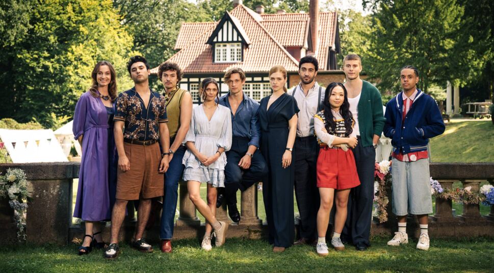 Auf dem Bild zum Cast von "Maxton Hall" Staffel 3 stehen zehn junge Schauspielerinnen und Schauspieler nebeneinander im Garten vor einem eleganten Landhaus mit rotem Ziegeldach. Die Stimmung wirkt freundlich und sommerlich. Alle sind modern und stilvoll gekleidet, was auf eine Mischung aus Drama und jugendlicher Eleganz hinweist. Die Figuren posieren locker und entspannt.