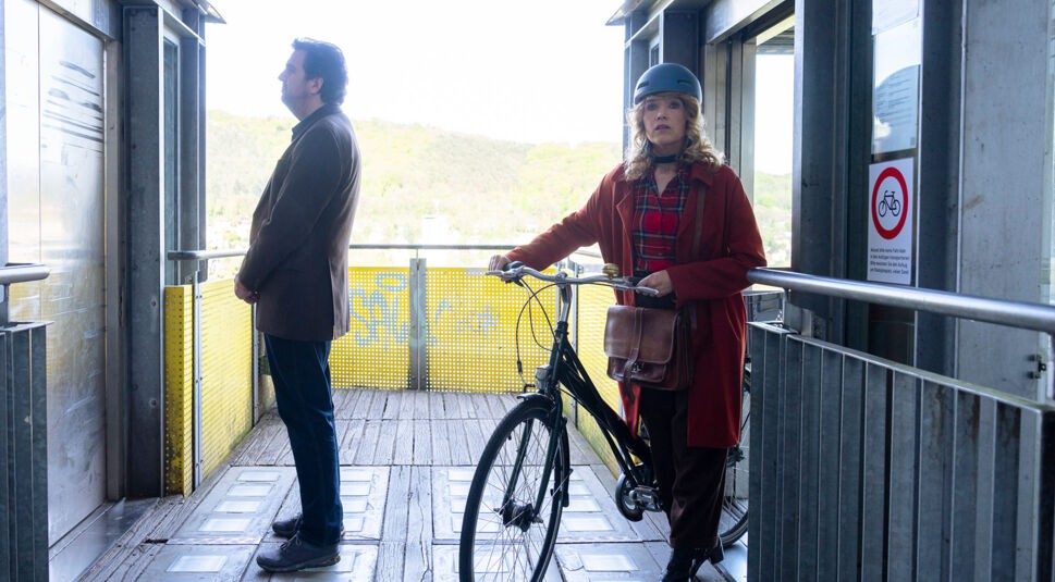 Auf dem Bild in dem Artikel zu Perfekt zusammen ist eine Szene aus der Prime-Video-Serie Perfekt verpasst zu sehen. Eine Frau und ein Mann stehen auf einer Plattform vor einem Aufzug. Die Frau trägt einen roten Mantel, einen Fahrradhelm und hält ein Fahrrad, während der Mann seitlich steht und ruhig wartet.