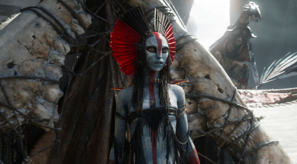 Auf dem Bild aus Avatar 3 ist eine weibliche Na’vi mit blauer Haut und auffälliger rot-schwarzer Kopfbedeckung zu sehen. Ihr Gesicht ist mit einem roten Streifen bemalt.