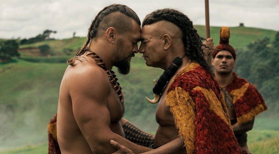 Auf dem Bild aus Chief of War sind Jason Momoa (links) und Temuera Morrison (rechts) in einer emotionalen Szene zu sehen. Beide Männer stehen Stirn an Stirn, eine Geste des Respekts und der Verbundenheit. Sie tragen traditionelle Kleidung und stehen in einer grünen Hügellandschaft. Die Szene wirkt würdevoll und bedeutungsvoll. 