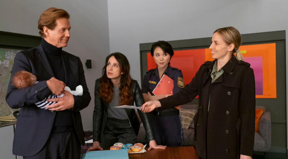 Das Artikelbild zu den Der Alte-Darsteller:innen zeigt Thomas Heinze als Hauptkommissar Caspar Bergmann, Stephanie Stumph als Kommissarin Annabell Lorenz, Yun Huang als IT-Spezialistin Julia Lulu Zhao und Annika Blendl als Richterin Eva Reinhardt (v.l.n.r.) zu sehen. Die Szene zeigt ein Gespräch im Büro, Heinze hält ein Baby, während Blendl ein Dokument überreicht.