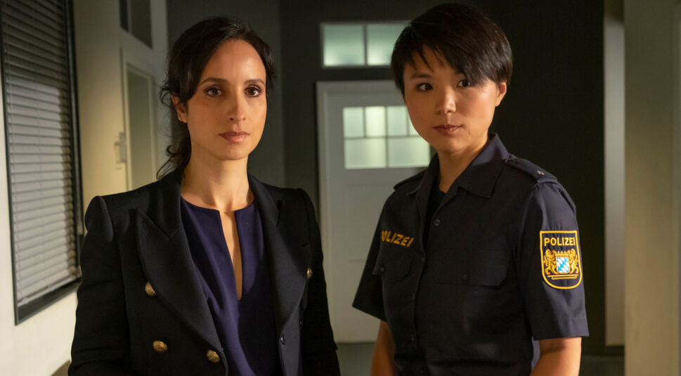 Auf dem Bild zu den Der Alte-Darsteller:innen sind Stephanie Stumph als Kommissarin Annabell Lorenz und Yun Huang als IT-Spezialistin Julia Lulu Zhao zu sehen. Stumph trägt ein dunkles Sakko mit goldenen Knöpfen, Huang eine Polizeiuniform mit bayerischem Wappen. Beide blicken ernst in die Kamera und stehen in einem Büroflur mit gedämpfter Beleuchtung.