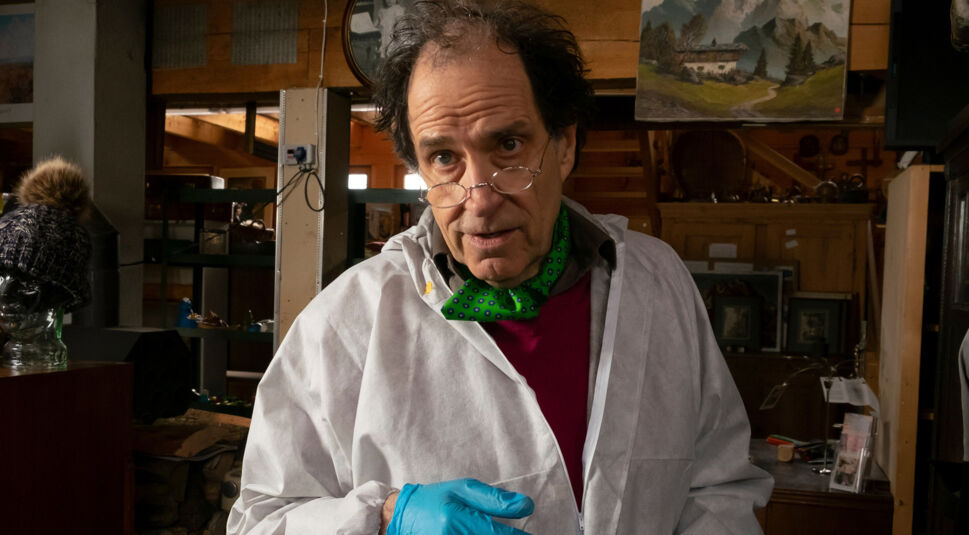 Auf dem Bild zur Der Alte Besetzung ist Helmfried von Lüttichau als Prof. Emerald Spindler zu sehen. Er trägt einen weißen Laborkittel, blaue Handschuhe und eine Brille, die tief auf seiner Nase sitzt. Sein Blick wirkt konzentriert und leicht kritisch. Die Umgebung erinnert an ein Atelier oder Labor, mit Holzinterieur und verschiedenen Gegenständen im Hintergrund, was eine investigative, leicht angespannte Stimmung erzeugt.