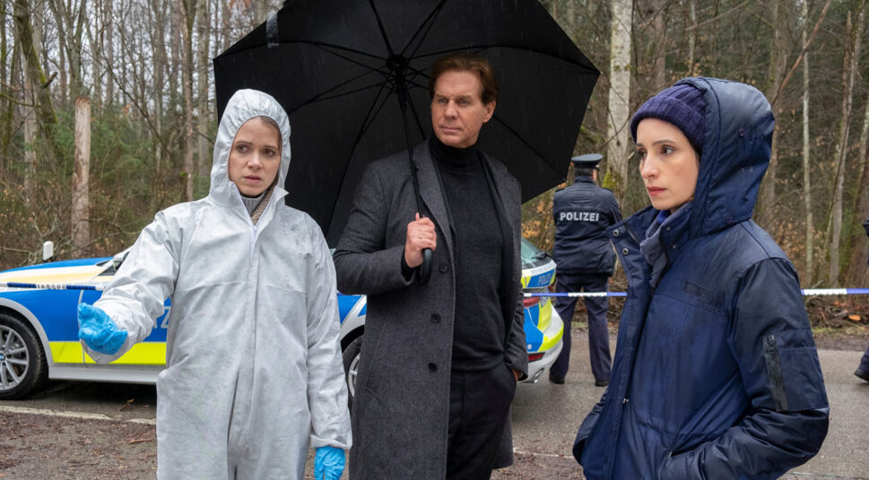 Auf dem Artikelbild zu den Der Alte-Darsteller:innen stehen Sidonie von Krosigk als Gerichtsmedizinerin Dr. Luisa Geiger, Thomas Heinze als Hauptkommissar Caspar Bergmann und Stephanie Stumph als Kommissarin Annabell Lorenz (v.l.n.r.) an einem regnerischen Tatort im Wald. Heinze hält einen schwarzen Regenschirm, während Krosigk einen weißen Ganzkörperanzug und blaue Handschuhe trägt.