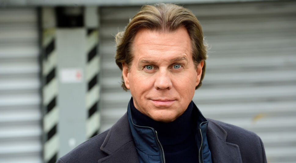 Auf dem Bild zu den Der Alte-Darsteller ist Thomas Heinze als Hauptkommissar Caspar Bergmann in einer Nahaufnahme zu sehen. Er trägt einen dunklen Rollkragenpullover, darüber eine gesteppte Jacke und einen Mantel. Heinze blickt direkt in die Kamera, im Hintergrund ist eine graue Rolltür mit schwarz-weißen Warnstreifen zu erkennen.