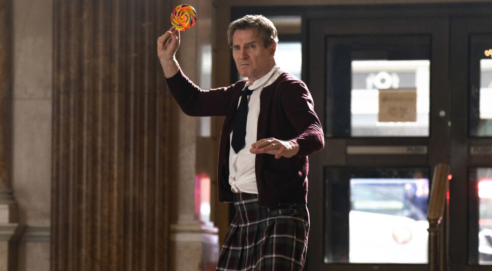 Liam Neeson ist in der Rolle des Frank Drebin Jr. in einer absurden Actionszene aus dem neuen Die nackte Kanone Film zu sehen. In angespannter Kampfpose hält er einen riesigen Lolli wie eine Waffe. Er trägt ein zerzaustes Hemd, Krawatte, Strickjacke und einen karierten Kilt – eine typische Mischung aus Slapstick und Ernst, wie sie für Die nackte Kanone charakteristisch ist.
