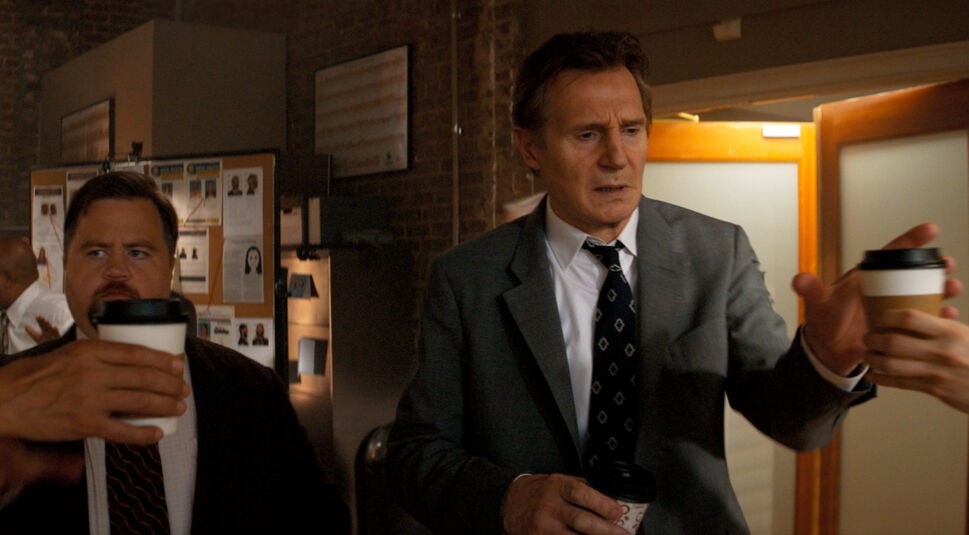 In einer Szene aus Die nackte Kanone sind Paul Walter Hauser als Ed Hocken Jr. und Liam Neeson als Frank Drebin Jr. in einem hektischen Büro zu sehen. Beide tragen Anzug und halten Kaffeebecher, während sie sich angespannt durch den Raum bewegen. Die Szene vermittelt eine Mischung aus Chaos und Komik – typisch für Die nackte Kanone.