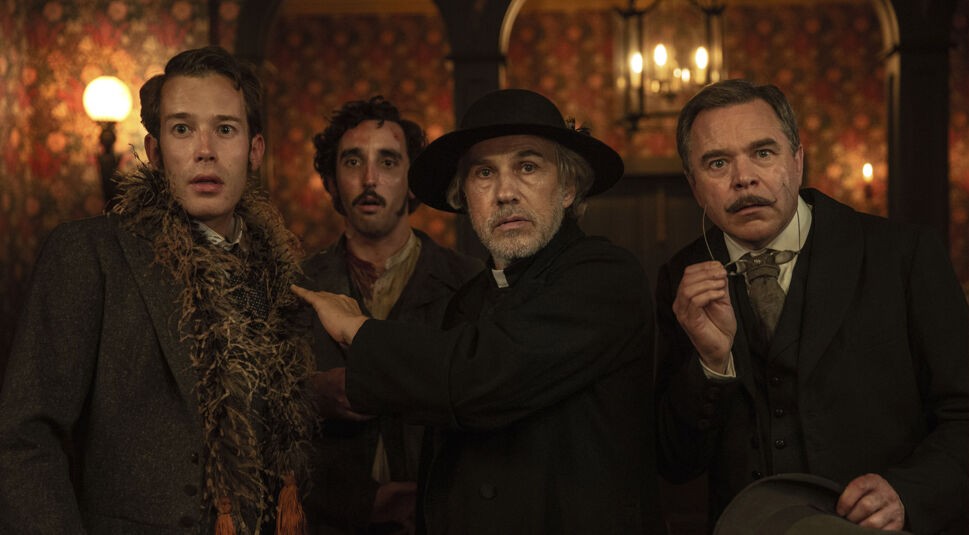 In dieser Szene aus „Dracula - Die Auferstehung“ steht der Priester, gespielt von Christoph Waltz, im Zentrum und blickt mit ernster Miene nach vorn. Um ihn herum stehen drei weitere Männer in viktorianischer Kleidung, alle mit erschrockenen oder angespannten Gesichtsausdrücken. Der opulente, warm beleuchtete Innenraum verstärkt die dramatische Spannung der Szene.