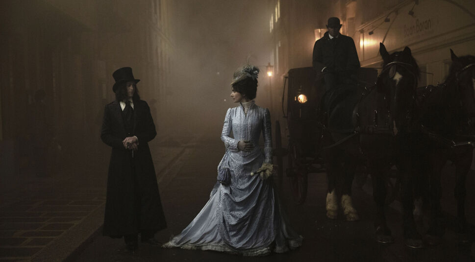 In dieser Szene aus Dracula - Die Auferstehung stehen Mina, gespielt von Zoe Bleu, und Dracula, gespielt von Caleb Landry Jones, in einer düsteren, nebelverhangenen Straße des viktorianischen Londons. Mina trägt ein elegantes blaues Kleid und blickt zurückhaltend zu Dracula, der in einem langen schwarzen Mantel und Zylinder bedrohlich wirkt. Eine Pferdekutsche verstärkt die düstere Atmosphäre.