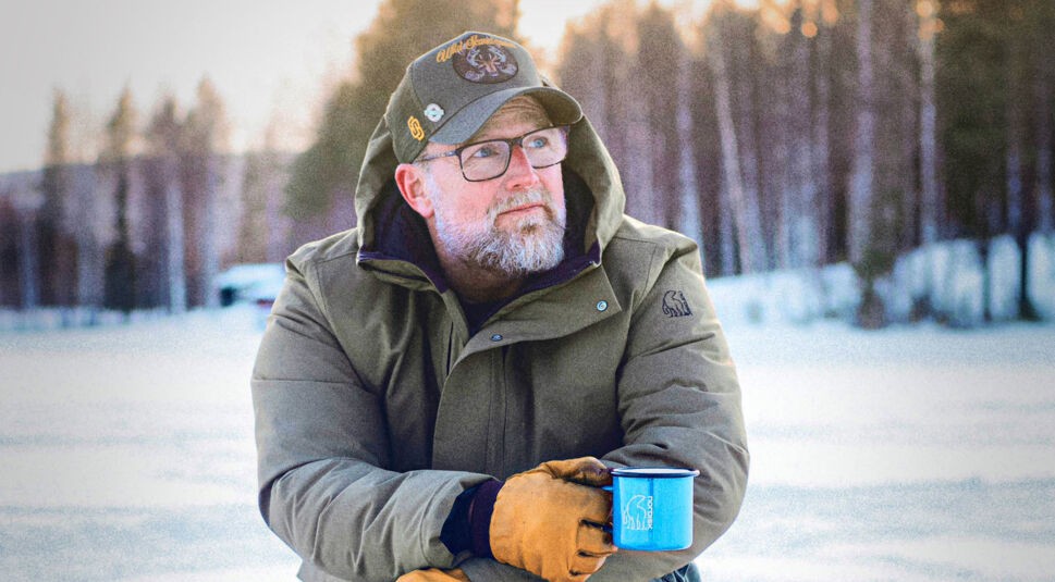 Auf dem Bild zur GO Real Life-Kampagne ist Brian Bojsen in winterlicher Umgebung zu sehen. Er trägt eine grüne Outdoorjacke mit Nordisk-Logo, eine Baseballkappe und Handschuhe. In der Hand hält er eine blaue Emaille-Tasse von Nordisk. Der Blick in die Ferne wirkt ruhig und reflektiert – die Szene strahlt Naturverbundenheit und Gelassenheit aus.