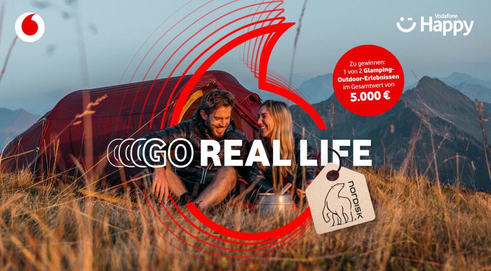 In dieser Szene der Vodafone-Kampagne „GO Real Life“ sitzt ein junges Paar lachend vor einem roten Nordisk-Zelt inmitten einer Berglandschaft. Die warme Abendstimmung unterstreicht das Naturerlebnis. Die „GO Real Life“-Werbung wirbt für ein Glamping-Gewinnspiel im Wert von 5.000 € und zeigt Naturverbundenheit und Lebensfreude.