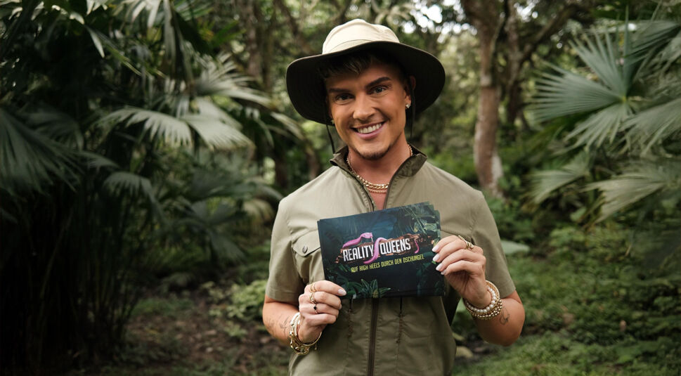 Auf dem Bild zu Reality Queens Staffel 2 steht der Influencer Twenty4Tim vor Bäumen. Er trägt ein Safari-Outfit mit breitem Hut und hält lächelnd eine Karte mit dem Logo der Sendung "Reality Queens – Auf High Heels durch den Dschungel" in der Hand.