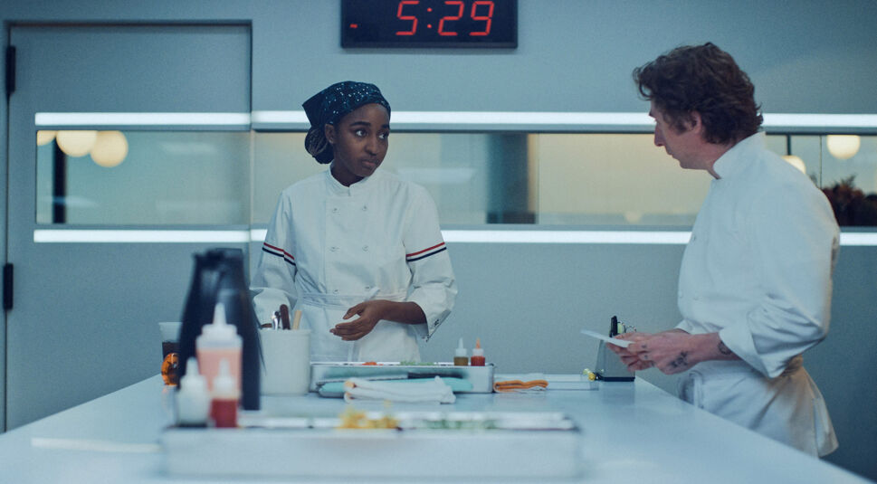 Auf dem Bild in dem Artikel zu einer möglichen The Bear Staffel 5 sind Syd und Carmy zu sehen. Beide tragen weiße Kochjacken und befinden sich in einer Küche. Syd blickt ernsthaft und aufmerksam zu Carmy, der einen Zettel in der Hand hält.