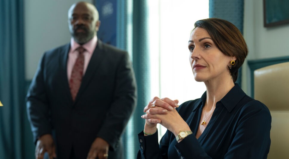 Suranne Jones in Hostage: Eine Frau sitzt mit verschränkten Händen und fokussiertem Blick an einem Schreibtisch, während im Hintergrund ein Mann im Anzug steht.