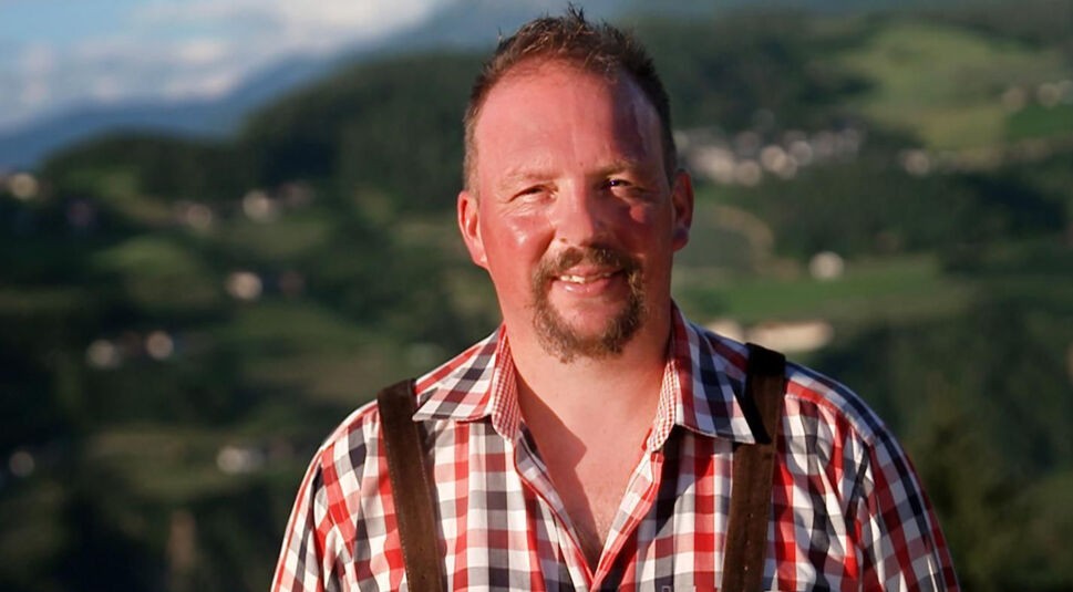 Auf dem Bild zu Bauer sucht Frau International 2026 steht ein Landwirt mit kariertem Hemd in den Bergen und blickt lächelnd in die Kamera.