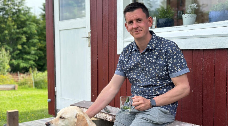 Auf dem Bild zu Bauer sucht Frau International 2026 sitzt ein Mann entspannt vor einem roten Holzhaus, hält ein Glas in der Hand und streichelt einen Hund.