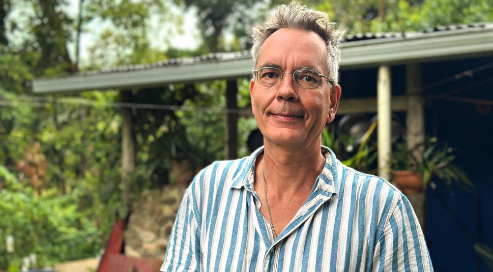 Auf dem Bild zu Bauer sucht Frau International 2026 steht ein Mann mit gestreiftem Hemd und Brille vor einem Haus mit Garten.