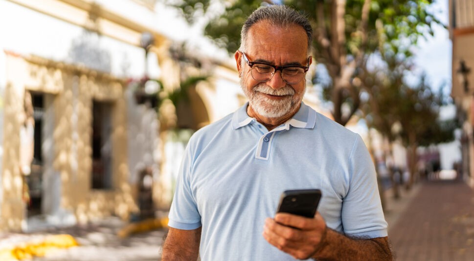 Auf dem Bild in dem Artikel zu den besten Handytarifen für Senior:innen steht ein älterer Mann mit Brille und grauem Bart im Sonnenlicht auf einer Straße. Er trägt ein hellblaues Polohemd und blickt zufrieden auf sein Smartphone.