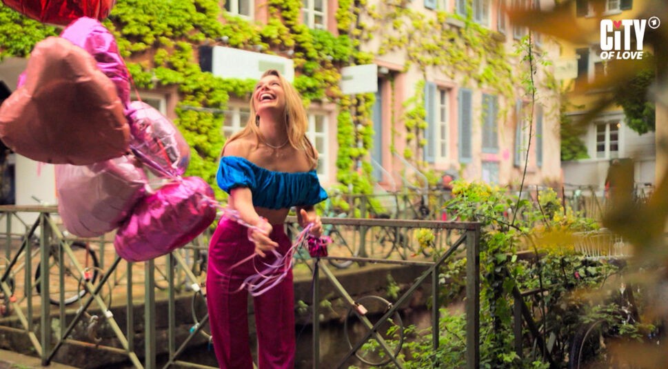 Auf dem Bild in dem Artikel zur ARD-Dating-Show City of Love zieht Lola Weippert mit fröhlichem Lachen mehrere herzförmige Luftballons durch die Luft. Sie steht auf einer kleinen Brücke vor einem Haus mit bewachsenen Fassaden und trägt ein blaues Oberteil sowie eine pinke Hose.