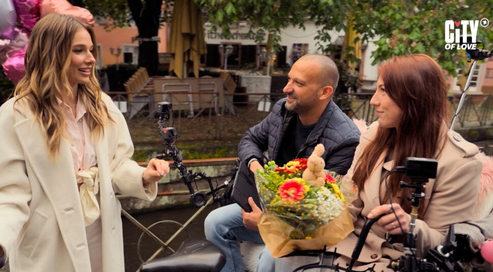 Auf dem Bild in dem Artikel zur ARD-Dating-Show City of Love spricht Lola Weippert mit zwei Kandidat:innen. Sie trägt einen hellen Mantel und lächelt, während die beiden Kandidat:innen auf einer Rikscha mit einem Blumenstrauß sitzen.