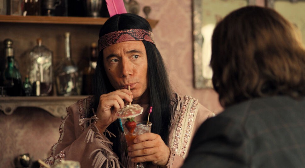 Auf dem Bild zur Das Kanu des Manitu Kritik ist Michael Bully Herbig in seiner Rolle als Winnetouch zu sehen. Er trägt ein glitzernd verziertes Outfit mit pinker Feder und trinkt genüsslich einen Cocktail mit Schirmchen und Erdbeere. Sein Blick ist aufmerksam, während ihm eine Person mit langen Haaren gegenübersitzt. Die Szene wirkt humorvoll und leicht ironisch.