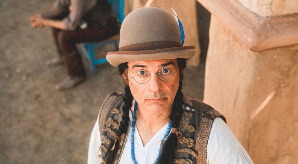 Auf dem Bild zur Das Kanu des Manitu Kritik ist Rick Kavanian in seiner Rolle als Dimitri zu sehen. Er blickt mit ernster Miene und großen Augen direkt in die Kamera. Dimitri trägt eine runde Brille, einen Filzhut mit Feder sowie zwei geflochtene Zöpfe. Im Hintergrund sitzt unscharf eine weitere Person auf einem blauen Stuhl.