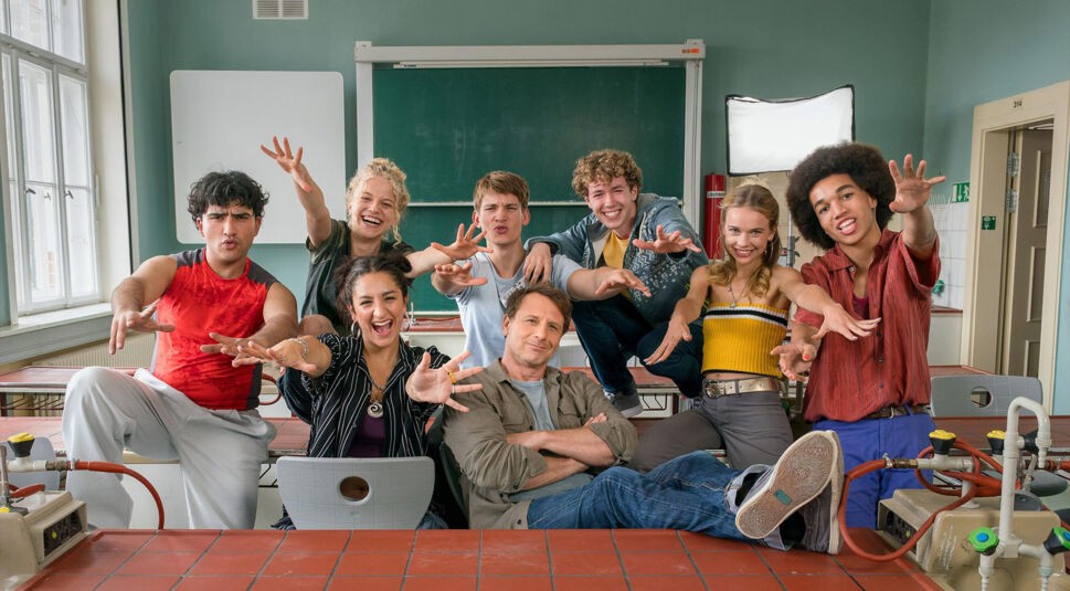 Auf dem Bild zur Serie Der Lehrer bei RTL sind acht Jugendliche und ein Lehrer in einem Klassenzimmer zu sehen. Sie posieren vor einer Schultafel, einige sitzen auf Tischen, andere stehen dahinter.