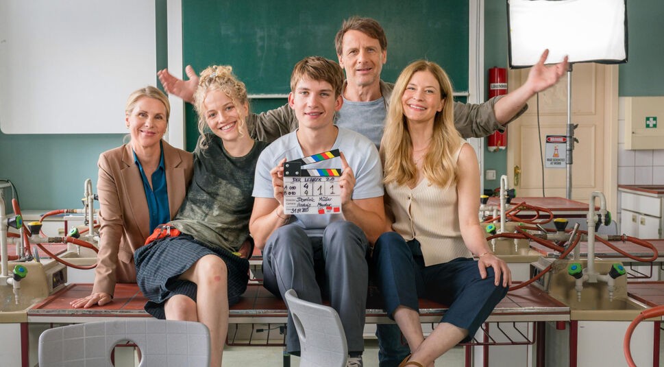 Dieses Bild zur Serie Der Lehrer bei RTL zeigt fünf Personen, die lächelnd auf einem Tisch im Klassenzimmer sitzen. In der Mitte hält eine junge Person eine Filmklappe mit dem Titel „Der Lehrer 2.0“.