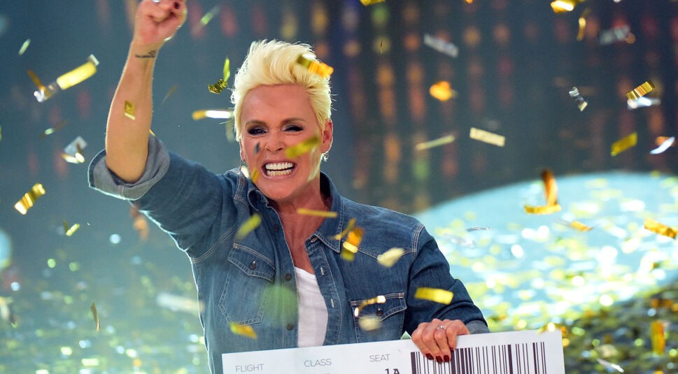 Die Dschungelcamp-Gewinnerin Brigitte Nielsen jubelt auf dem Bild unter einem goldenen Konfettiregen. Sie trägt ein Jeanshemd, hält stolz ein übergroßes Ticket in der Hand und strahlt mit erhobener Faust in die Kamera. Ihr platinblondes, nach hinten gestyltes Haar und ihre energiegeladene Pose unterstreichen den Moment des Sieges auf der Showbühne.