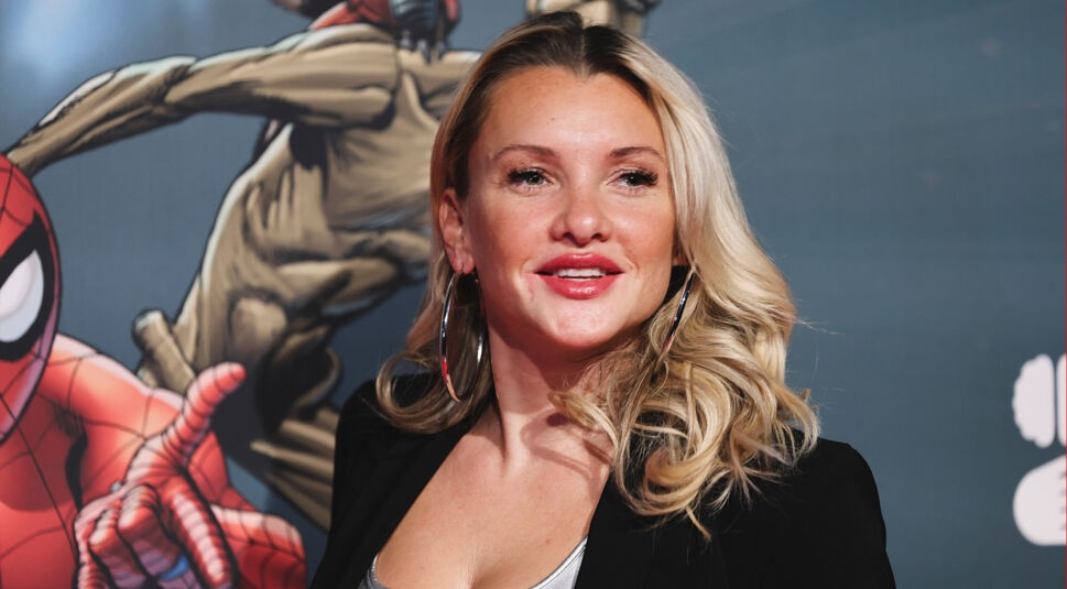 Die Dschungelcamp-Gewinnerin Evelyn Burdecki steht lächelnd vor einer Comic-Wand mit Spider-Man-Motiv. Sie trägt große silberne Creolen, ein schwarzes Jackett und ein silberfarbenes Top. Ihre blonden, gewellten Haare fallen locker über die Schultern. Die Szene wirkt lebendig und glamourös, passend zu einem Popkultur-Event.