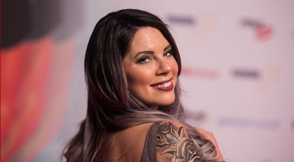 Auf dem Bild ist die Dschungelcamp-Gewinnerin Jenny Frankhauser zu sehen. Sie blickt lächelnd über die Schulter in die Kamera und trägt ein schulterfreies Outfit, das ein großes Rosen-Tattoo auf ihrem Oberarm zeigt. Ihre langen, gewellten Haare verlaufen in einem dunklen bis lilafarbenen Ton. Der Hintergrund zeigt eine Veranstaltung mit Logos und Sponsorenwänden.