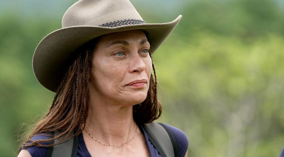 Auf dem Bild ist Dschungelcamp-Gewinnerin Lilly Becker in einem natürlichen Dschungel-Setting zu sehen. Sie trägt einen breitkrempigen Hut und schaut ernst, aber gefasst in die Ferne. Ihr Look ist natürlich, mit geflochtenen Haarsträhnen und einem Rucksack auf den Schultern – bereit für das Abenteuer. Der grüne, verschwommene Hintergrund unterstreicht die Wildnis-Stimmung.
