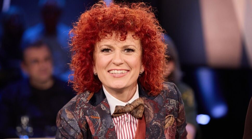 Auf dem Bild ist die Dschungelcamp-Gewinnerin Lucy Diakovska zu sehen. Sie sitzt in einem Studio und lächelt direkt in die Kamera. Lucy trägt ein auffälliges, florales Sakko mit Fliege und gestreiftem Hemd. Ihre roten, lockigen Haare und ihre fröhliche Ausstrahlung stechen hervor. Die Studiobeleuchtung sorgt für eine warme, einladende Atmosphäre.