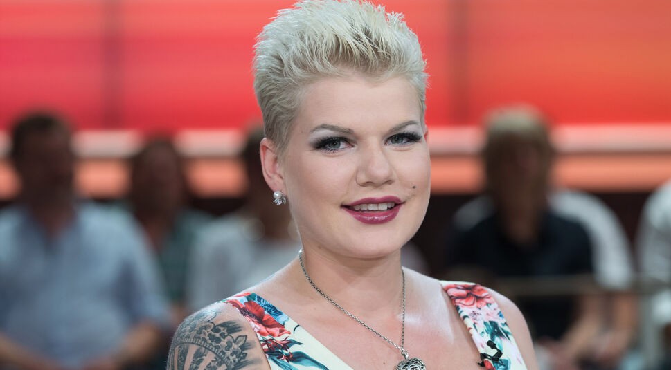 Auf dem Bild ist die Dschungelcamp-Gewinnerin Melanie Müller zu sehen. Sie trägt ein sommerliches, geblümtes Kleid und eine auffällige silberne Herzkette. Ihr blondes, kurzes Haar ist nach hinten gestylt, und ein großes Tattoo ziert ihren rechten Oberarm. Im Hintergrund sitzt ein unscharfes Publikum, die Stimmung wirkt freundlich und entspannt.