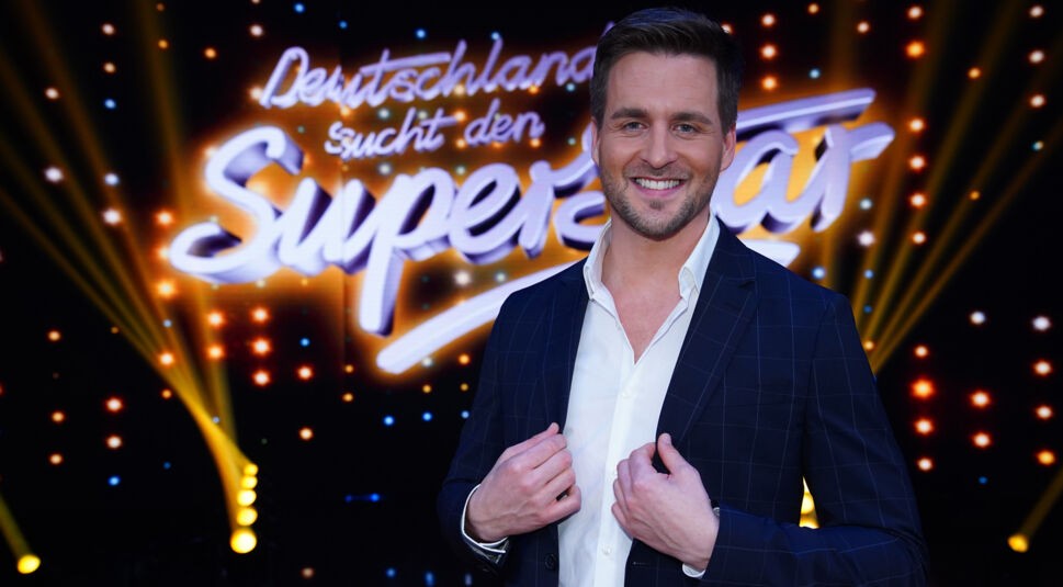 Auf dem Bild ist DSDS-Gewinner Alexander Klaws vor dem leuchtenden „Deutschland sucht den Superstar“-Logo zu sehen. Er trägt ein dunkles Sakko über einem weißen Hemd und lächelt selbstbewusst in die Kamera. Die festliche Studioatmosphäre mit Scheinwerfern und Sternenlicht unterstreicht seine Rolle als einer der bekanntesten DSDS-Gewinner:innen.