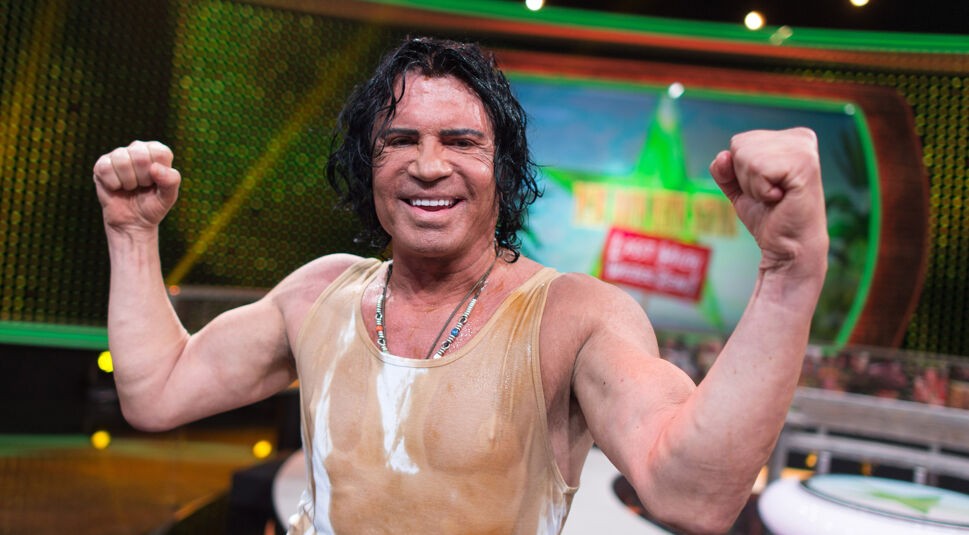 Auf dem Bild ist der Dschungelcamp-Gewinner Costa Cordalis zu sehen. Er steht auf einer Studiobühne, trägt ein verschwitztes, eng anliegendes Tanktop und zeigt beide Arme muskulös und triumphierend in Siegerpose. Sein Gesicht wirkt stolz und zufrieden, im Hintergrund ist das Logo der Dschungelshow zu erkennen. Die Stimmung ist feierlich und energiegeladen.