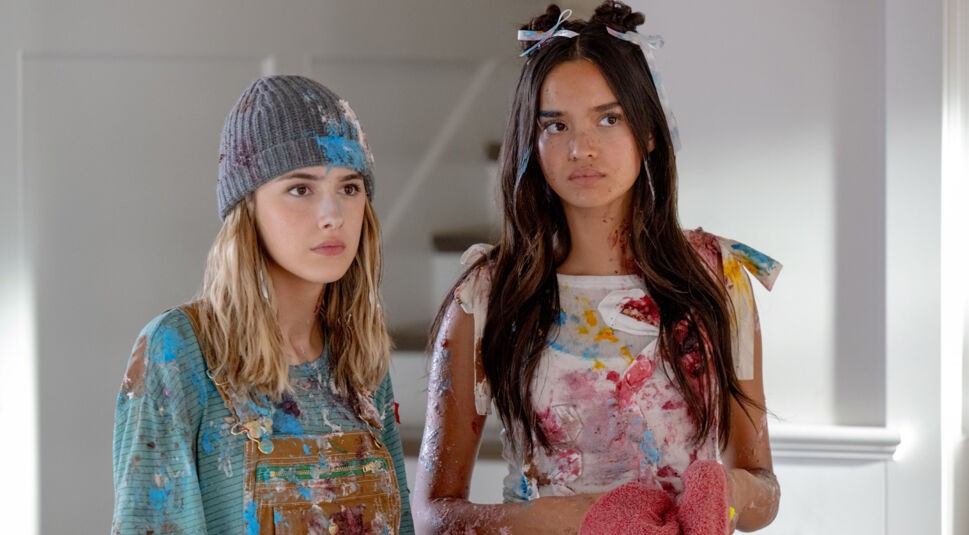 Im Bild aus Freakier Friday stehen Julia Butters als Harper Coleman und Sophia Hammons als Lily Davies nebeneinander in einem hellen Raum. Beide sind von Kopf bis Fuß mit bunter Farbe verschmiert und schauen ernst oder leicht genervt. Ihre Kleidung und Gesichtsausdrücke deuten auf ein kürzliches Chaos hin – eine humorvolle Szene für die Freakier Friday Kritik.