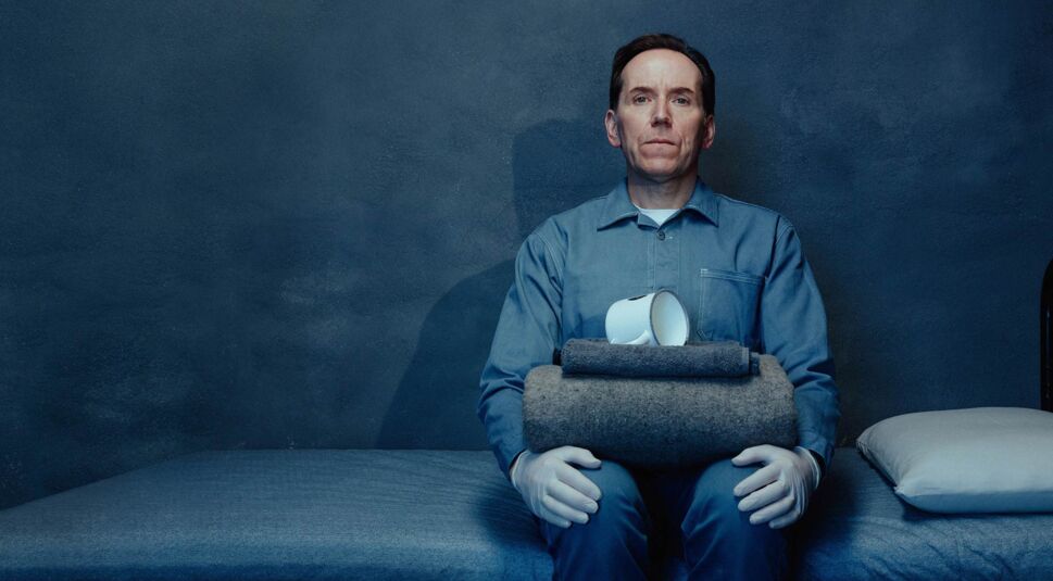 Das Bild in den GigaTV-Highlights im September 2025 zeigt Ben Miller als Professor T in der gleichnamigen Serie Professor T. In kühler, blauer Gefängniszelle sitzt er auf einem Bett, trägt Handschuhe und hält gefaltete Decken sowie eine umgestürzte Tasse.