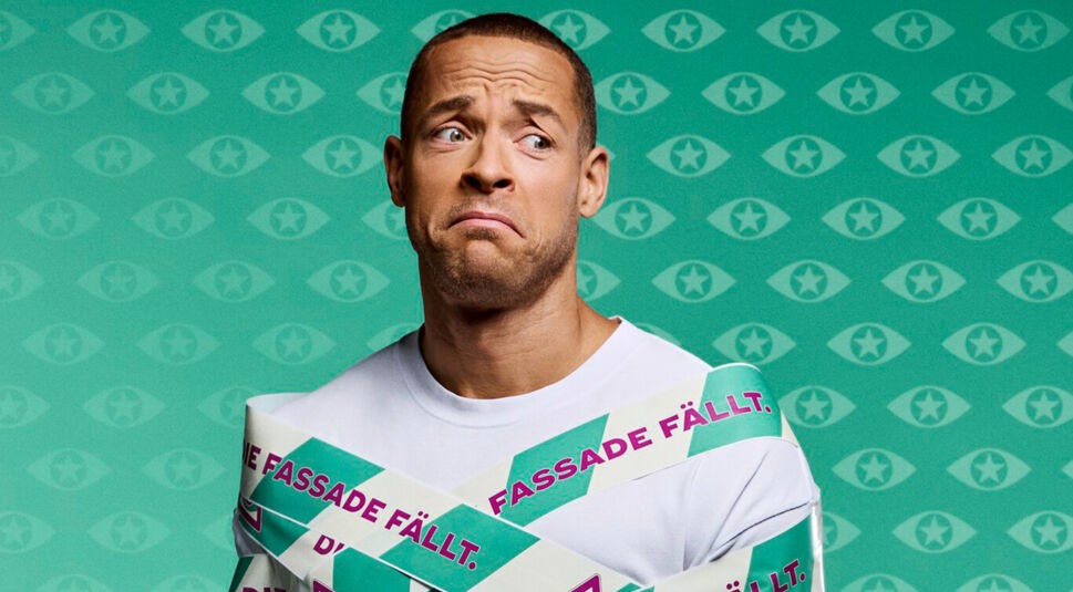 Auf dem Bild zur Sendung Promi Big Brother 2025 ist Andrej Mangold zu sehen, der mit Band umwickelt ist, auf dem „Die Fassade fällt.“ steht. Er trägt ein weißes Shirt und blickt mit skeptischem Gesichtsausdruck zur Seite.