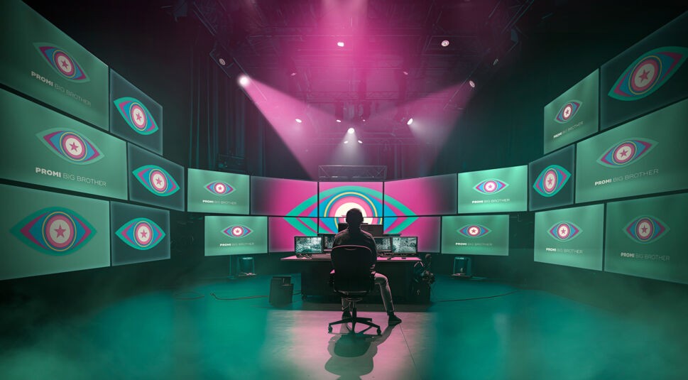 Das Bild zur Promi Big Brother 2025 zeigt einen Überwachungsraum mit zahlreichen Bildschirmen, auf denen das bekannte Big-Brother-Auge zu sehen ist. Im Zentrum sitzt eine Person im Schatten vor den Monitoren.