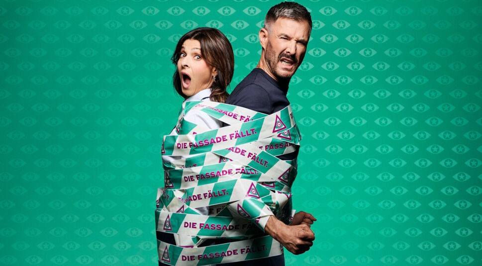 Auf dem Bild zur Show Promi Big Brother 2025 sind die Moderator:innen Marlene Lufen und Jochen Schropp zu sehen. Beide stehen Rücken an Rücken und sind mit auffälligem Klebeband umwickelt, auf dem „DIE FASSADE FÄLLT.“ steht. 